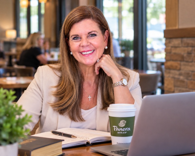 Carole Mendoza smiling at a café table with a Panera Bread coffee cup, an open notebook, a Bible and a laptop